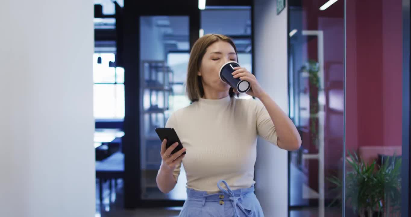 在现代办公室里拿着咖啡杯拿着智能手机走路的亚洲女商人视频素材