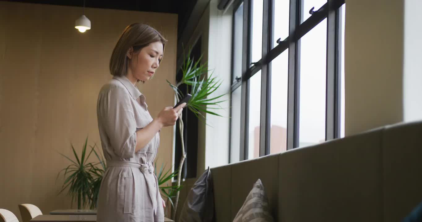 亚洲女商人站在窗口使用智能手机在现代办公室视频素材