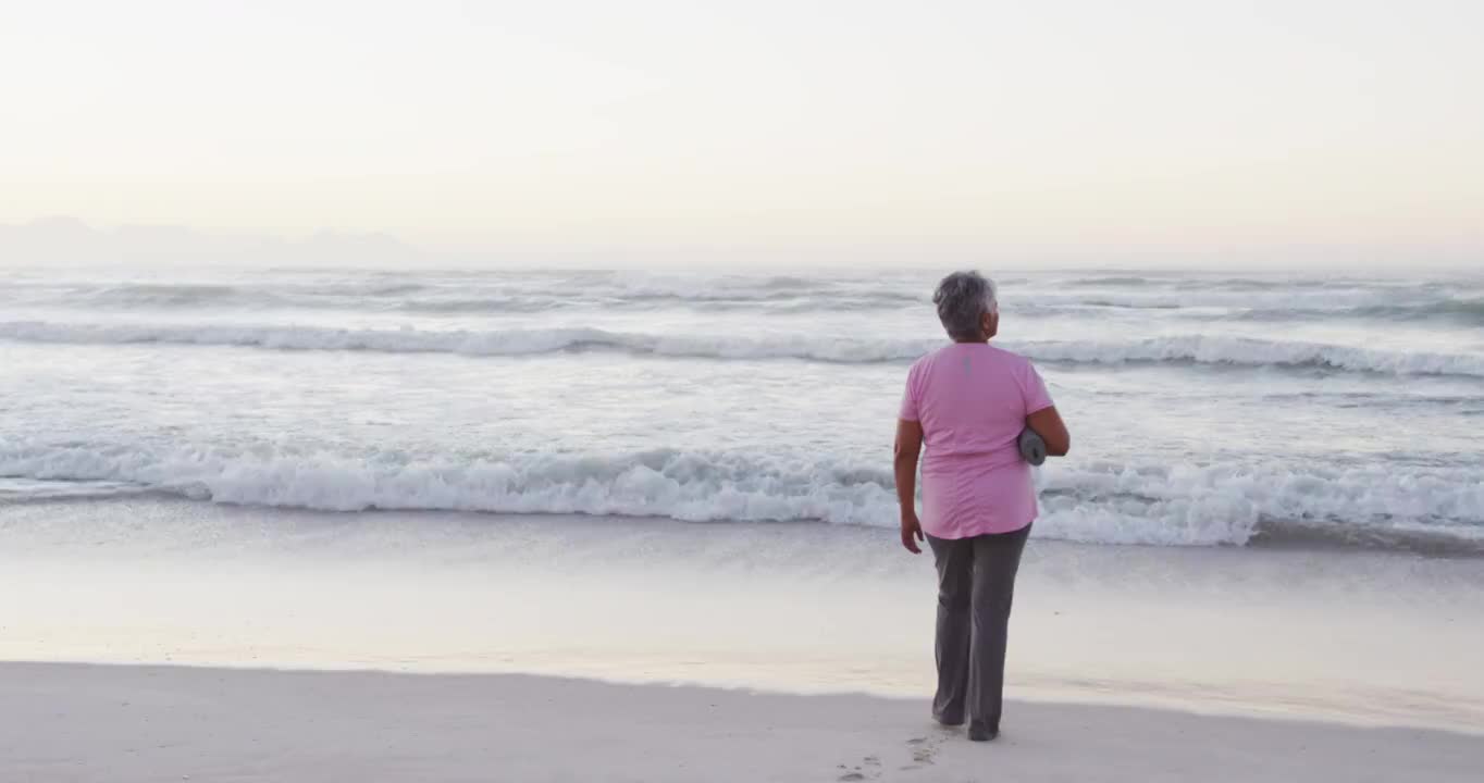 非裔美国高级妇女持有瑜伽垫看着海浪，而站在海滩上视频素材