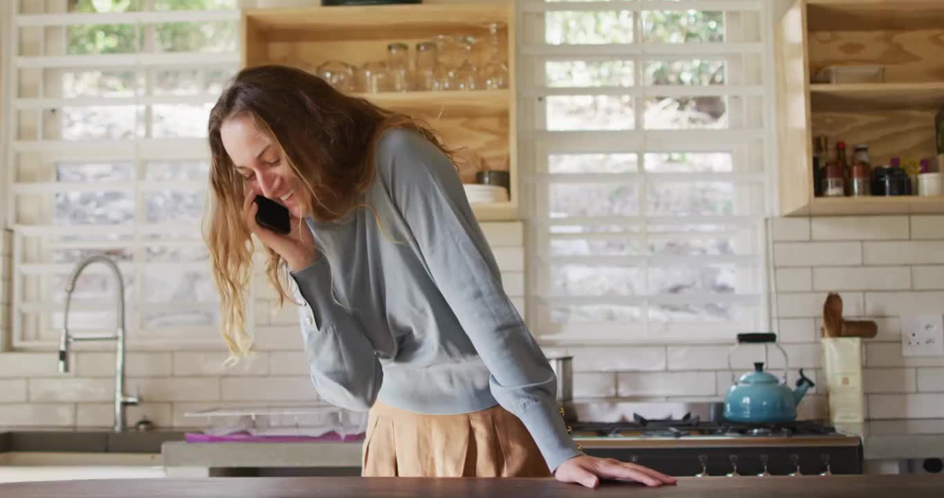 快乐的白人妇女站在阳光明媚的小屋厨房里用智能手机聊天，面带微笑视频素材