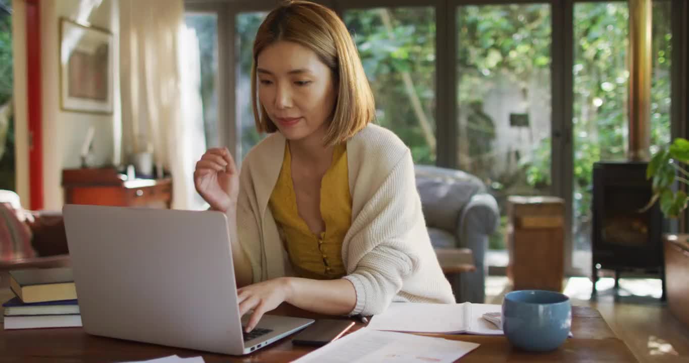 亚洲妇女坐在桌旁工作，从家里和使用笔记本电脑视频素材