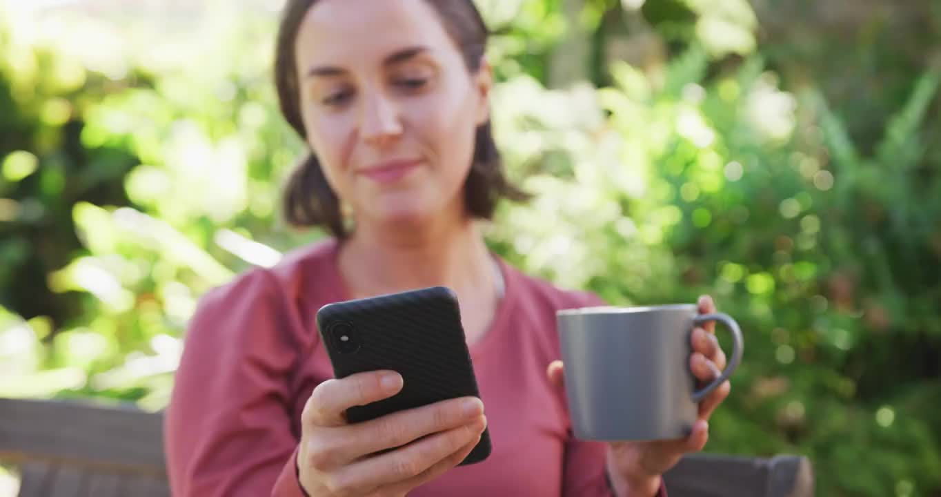 微笑的白人妇女使用智能手机，拿着杯茶坐在阳光花园视频素材