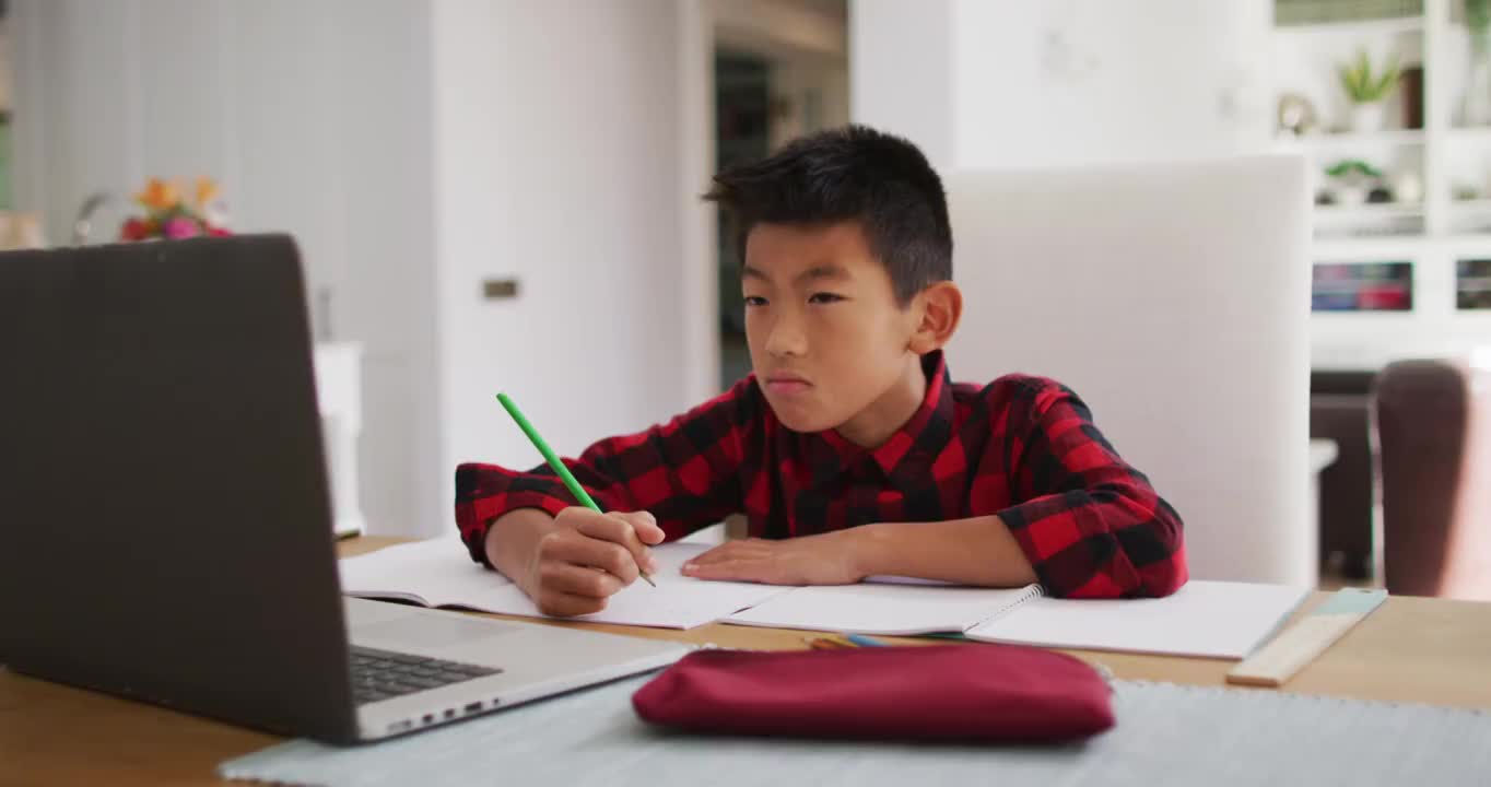 亚洲男孩在家，坐在桌旁使用笔记本电脑为在线学校课程视频素材