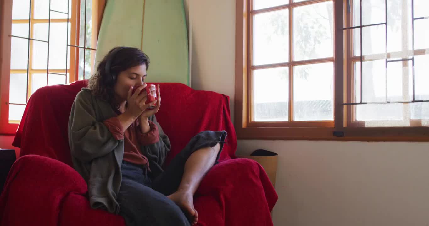 轻松的混血儿妇女戴着耳机坐在阳光明媚的小屋客厅喝茶视频素材