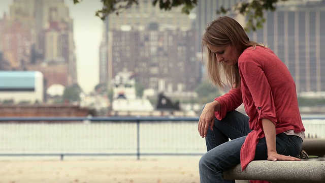 一个女人独自坐在公园的长椅上。当一艘驳船在哈德逊河上驶过时，她抬头看着纽约的天际线视频素材