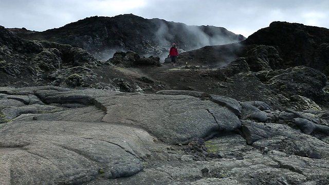 在冰岛Myvatn地区leirhnjukur / Krafla，蒸汽从黑色火山景观中倾泻而出，年轻女子走在火山岩上视频素材