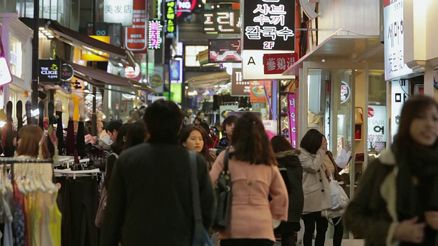 在韩国首尔的明洞中心购物区，购物者们在夜晚走过一条布满灯光招牌的繁忙街道视频素材