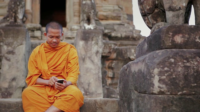 在柬埔寨暹粒吴哥窟(Angkor Wat / Siem Reap)的一座古庙上，一名和尚在用智能手机的屏幕上轻敲视频素材