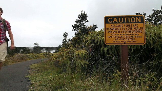 美国夏威夷火山国家公园/火山，大岛，夏威夷，一名年轻男子穿着登山服走向警示标志旁的小路，警示标志显示危险的火山烟雾视频素材