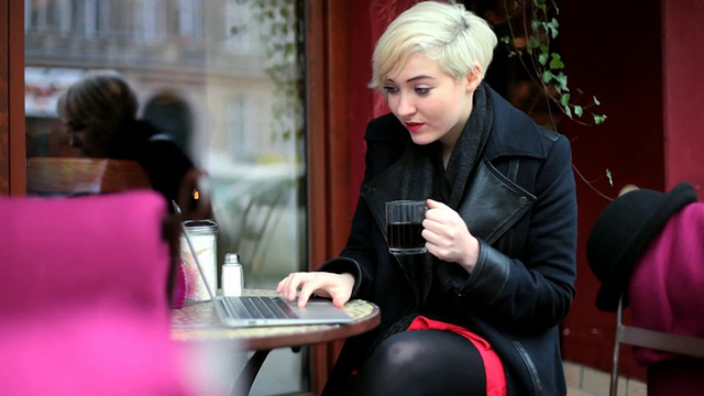 MS Shot of Woman drinking hot tea at cafe outside while talking on Skype and typing on laptop /柏林，德国视频素材