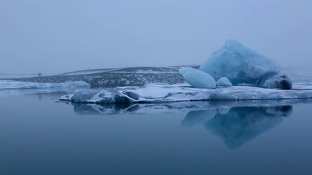 巨大的冰川漂浮在平静的Jokulsarlon泻湖的水面上，远处汽车驶过，年轻夫妇在冰岛湖边接吻视频素材