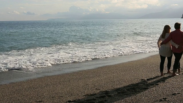 一对夫妇沿着海滩散步，海浪滚滚而来，注意了视频素材