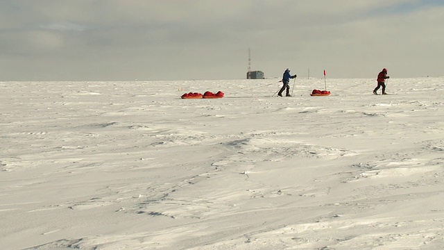 图为两个人在滑雪板上拉着雪橇穿过南极冰，上面有橙色标志旗和建筑/南极，南极洲视频素材