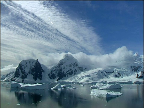 跟踪右上方令人惊叹的冰雪覆盖的山脉与小浮冰在前景的海洋和白色蓬松的云在天空南极洲视频素材