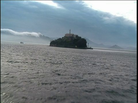 穿过平静的海面向远处的恶魔岛前进。阴沉的蓝色多云的天空和背景地平线倾斜的小山视频素材