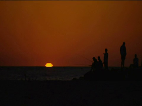 在佛罗里达海滩上，一群人在橙色的夕阳下钓鱼，太阳落下，消失在地平线后面视频素材