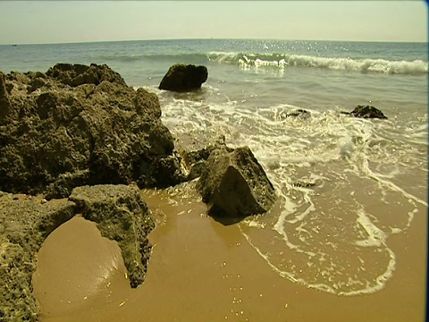 海浪拍打着岩石和沙滩，阳光反射出海面的背景。视频素材