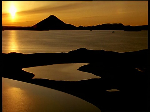 太阳在后面的山在背景反射金色的光表面平静的水，小泻湖在前景，冰岛视频素材