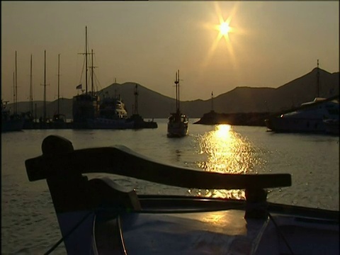 在夕阳西下的希腊，镜头从船舵上望向驶出港口的一艘船视频素材