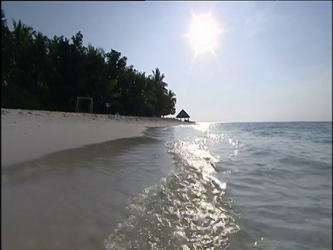 清澈的海水拍打着白色的沙滩，阳光反射在波光粼粼的海棕榈树和小海滩小屋的剪影映衬着明亮的蓝白色的天空在马尔代夫的背景视频素材