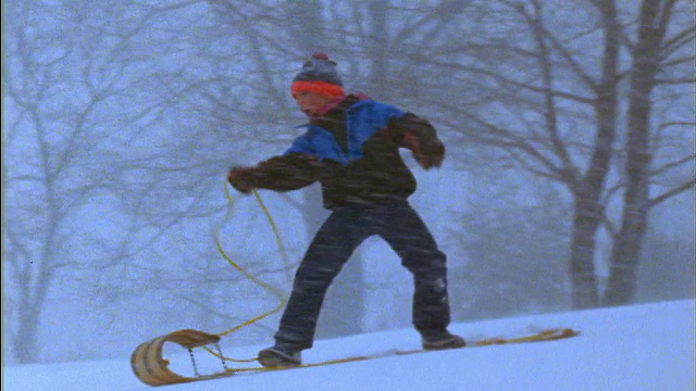 慢镜头:一个男孩在暴风雪中站在雪橇上下山+摔下来视频素材