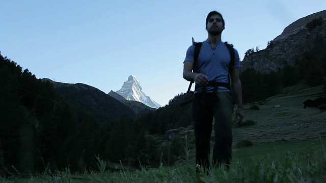 一个登山者走在绿色的田野上，背景是马特洪峰。视频素材