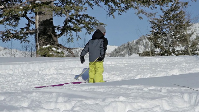 在一个阳光明媚的冬日，一个小男孩拉着他的雪橇穿过雪地视频素材