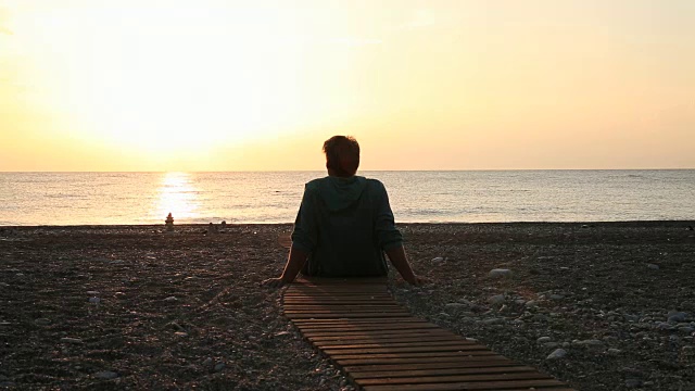 男子在海滩人行道上休息，看海面上的日出视频素材