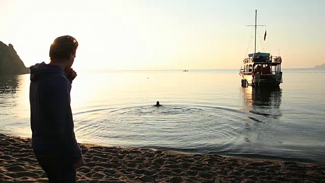 日出时分，男人在海滩边看着女人跳进海里视频素材