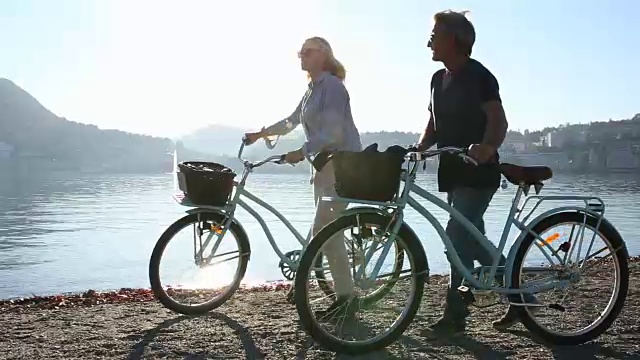 一对夫妇沿着湖边的海滩推着自行车，城市在后面视频素材