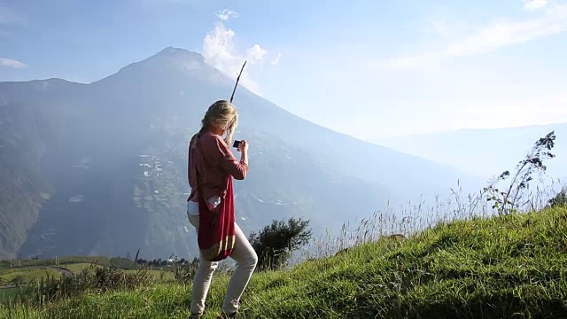 一名女子走过火山下的高山草地，用智能手机拍照视频素材