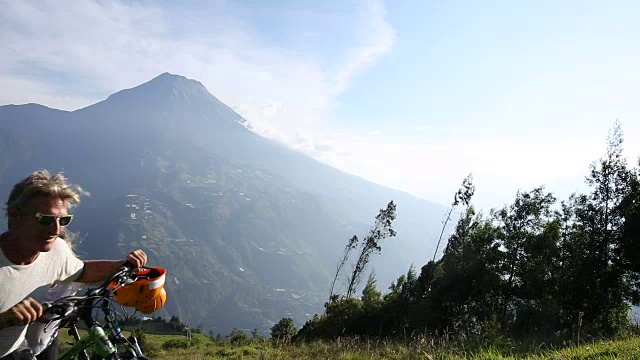 一对夫妇推着自行车穿过高山草地，后面是火山视频素材