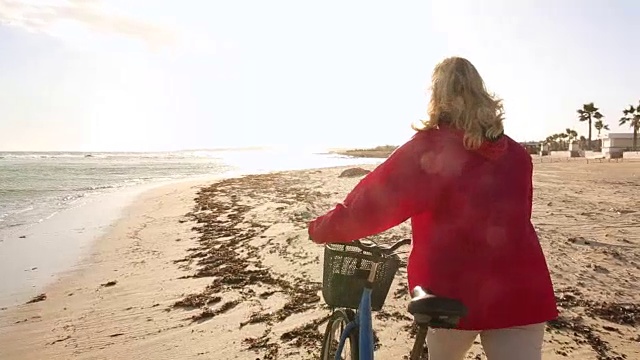 女人推着自行车沿着空荡荡的海滩，旁边是冲浪板视频素材