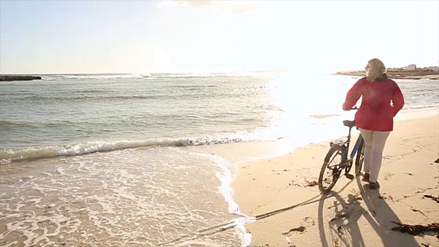 女人推着自行车沿着空荡荡的海滩，旁边是冲浪板视频素材