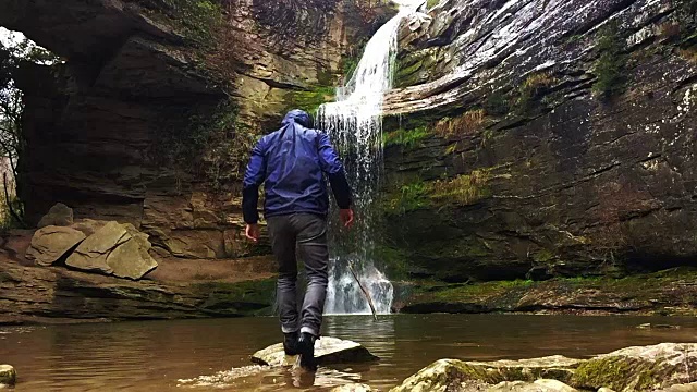 一个徒步旅行者在瀑布马尾溅在一个令人惊叹的风景与下雨的天气在一个公路旅行冒险山脉的慢动作视图。视频素材