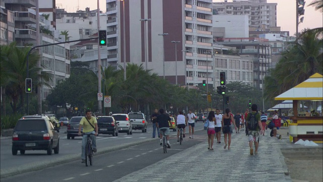 在巴西里约热内卢，骑自行车的人在一条繁忙的街道和伊帕内玛海滩之间的自行车道上骑行。视频素材