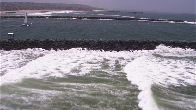 海浪冲刷着岩石码头，那里有几艘帆船在圣迭戈附近航行。视频素材