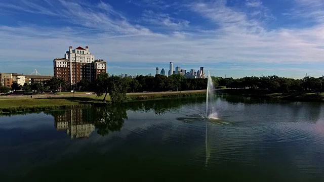 Aerial: Lake Cliff Park View flying close to water with amazing morning reflection with Fountain spraying high in the air and Dallas Texas Skyline in the background of Cityscape Turn .鸟瞰:湖崖公园近水楼台，早晨喷泉在空中喷溅，达拉斯德克萨斯天际线在城市景观的背景视频素材