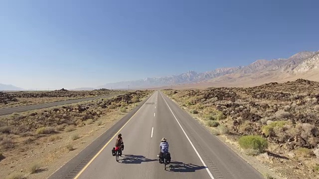 鸟瞰2名骑自行车的人沿着空旷的道路穿越沙漠视频素材