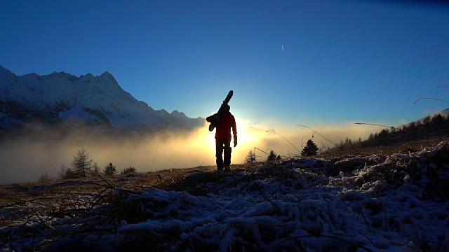 夕阳西下的雪山上，一个带着滑雪板的男人在行走视频素材