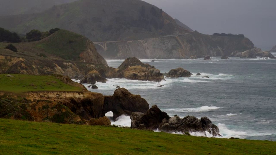 宽镜头的海洋坠毁在岩石海岸与山和桥在阴天的背景视频素材