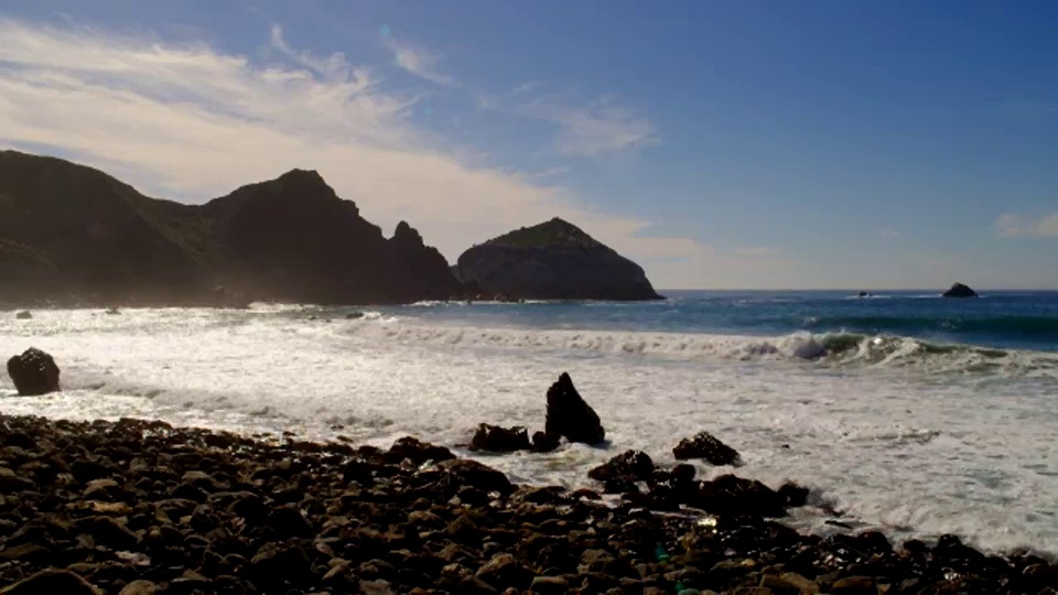 从中到宽镜头的海浪冲上岸岩石海滩与山脉在海岸的背景视频素材