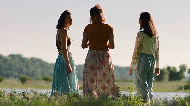 在阳光明媚的日子里，三个女孩站在河边的野花地里，指着天空。视频素材