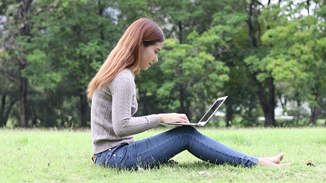 亚洲女孩微笑与笔记本电脑在户外公园/女孩笔记本电脑户外公园/亚洲女孩使用笔记本电脑在户外公园视频素材