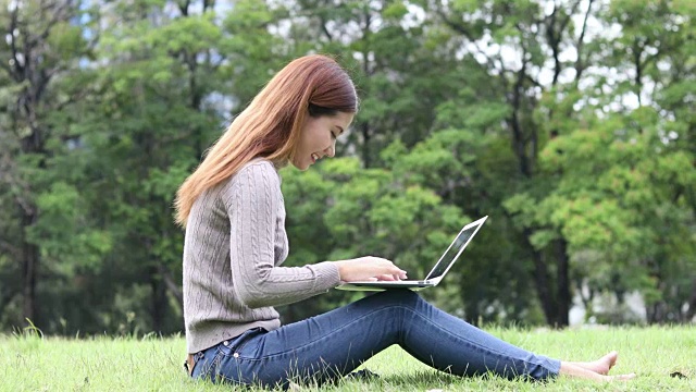 亚洲女孩微笑与笔记本电脑在户外公园/女孩笔记本电脑户外公园/亚洲女孩使用笔记本电脑在户外公园视频素材