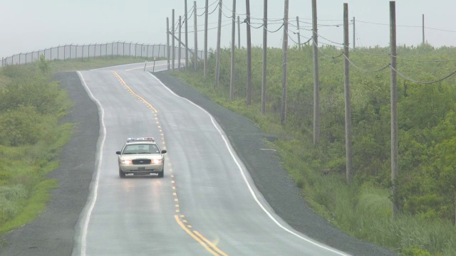 广角乡村道路。道路两旁可见围栏、电线杆和电线。汽车驾驶可见。警车可见。下雨了。视频素材