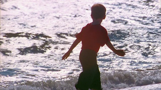 中等拍摄的小男孩站在海浪中，在海滩上捡起潮湿的沙子视频素材