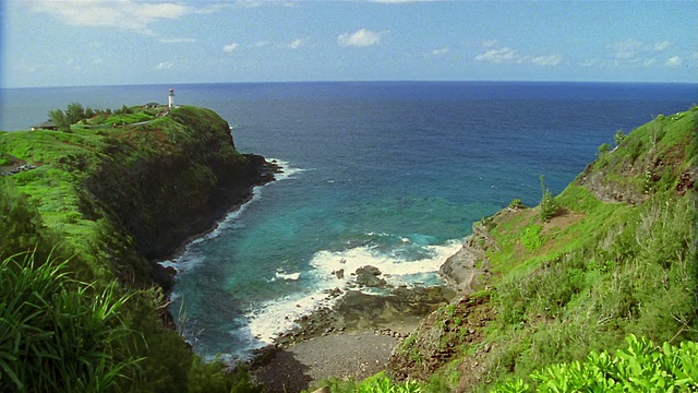 高角度宽镜头海浪撞击岩石海岸与灯塔在绿色悬崖/夏威夷视频素材