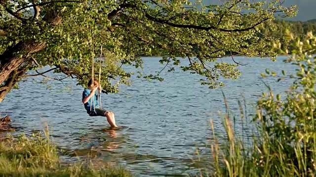 这是一个阳光明媚的夏日，夕阳西下，9岁的小男孩穿着牛仔裤、短裤和蓝色衬衫，坐在树上的秋千上，秋千悬挂在斯塔弗尔西湖的湖边，他厌倦了数码排毒，一边用脚在水里戏水视频素材