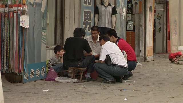 中国山东，男子服装店门前的人行道上，男人和男孩蜷缩在玩游戏的男人周围视频素材